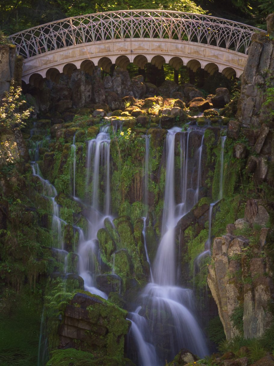 Ein Wasserfall fließt unter einer eleganten Brücke, umgeben von grünem Moos und Bäumen.