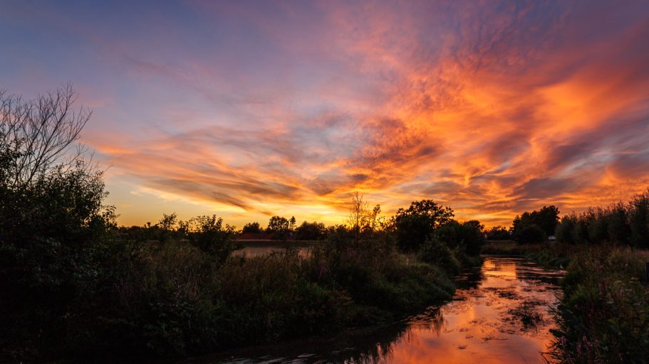 Dramatischer Sonnenuntergang über einem ruhigen Fluss, umgeben von Bäumen und Sträuchern.