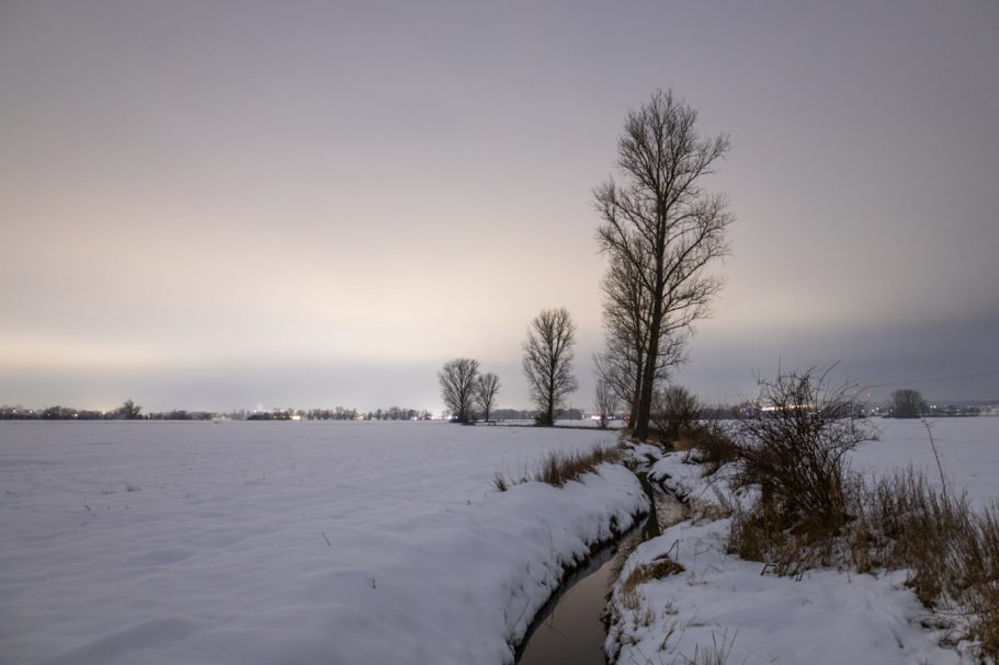 Schneebedecktes Feld mit Bäumen am Horizont und einem schmalen Wasserlauf.