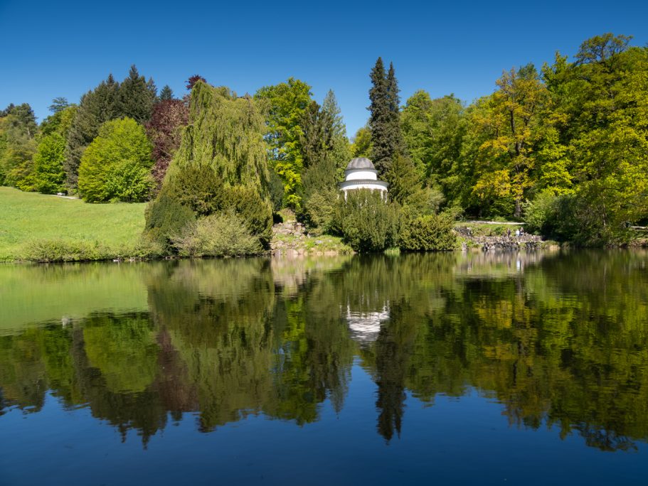 Ein ruhiger See spiegelt einen Pavillon und umgebende Bäume unter klarem Himmel.